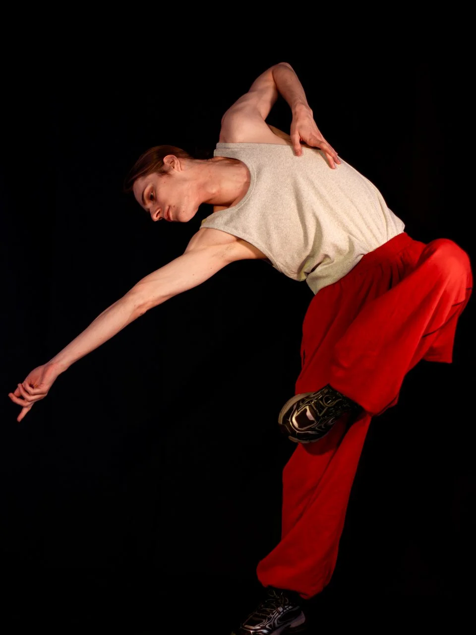A person with short hair in a gray sleeveless shirt and red pants performing a dance move against a black background, balancing on one leg with the other bent and reaching outward with one arm.
