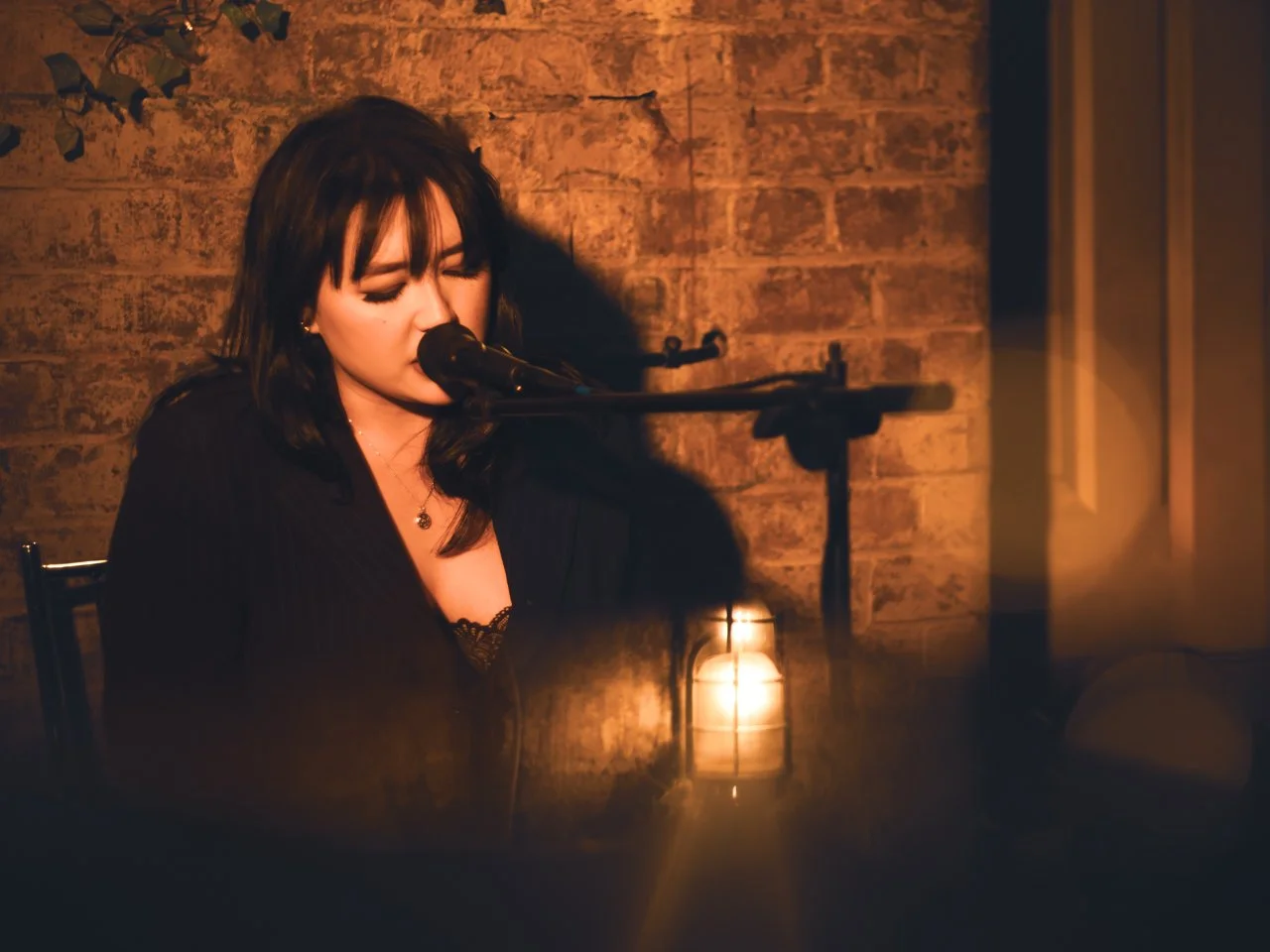 A woman with dark hair and bangs is singing into a microphone, seated at a small table with a lit candle, against a brick wall background in a cozy, dimly lit setting.