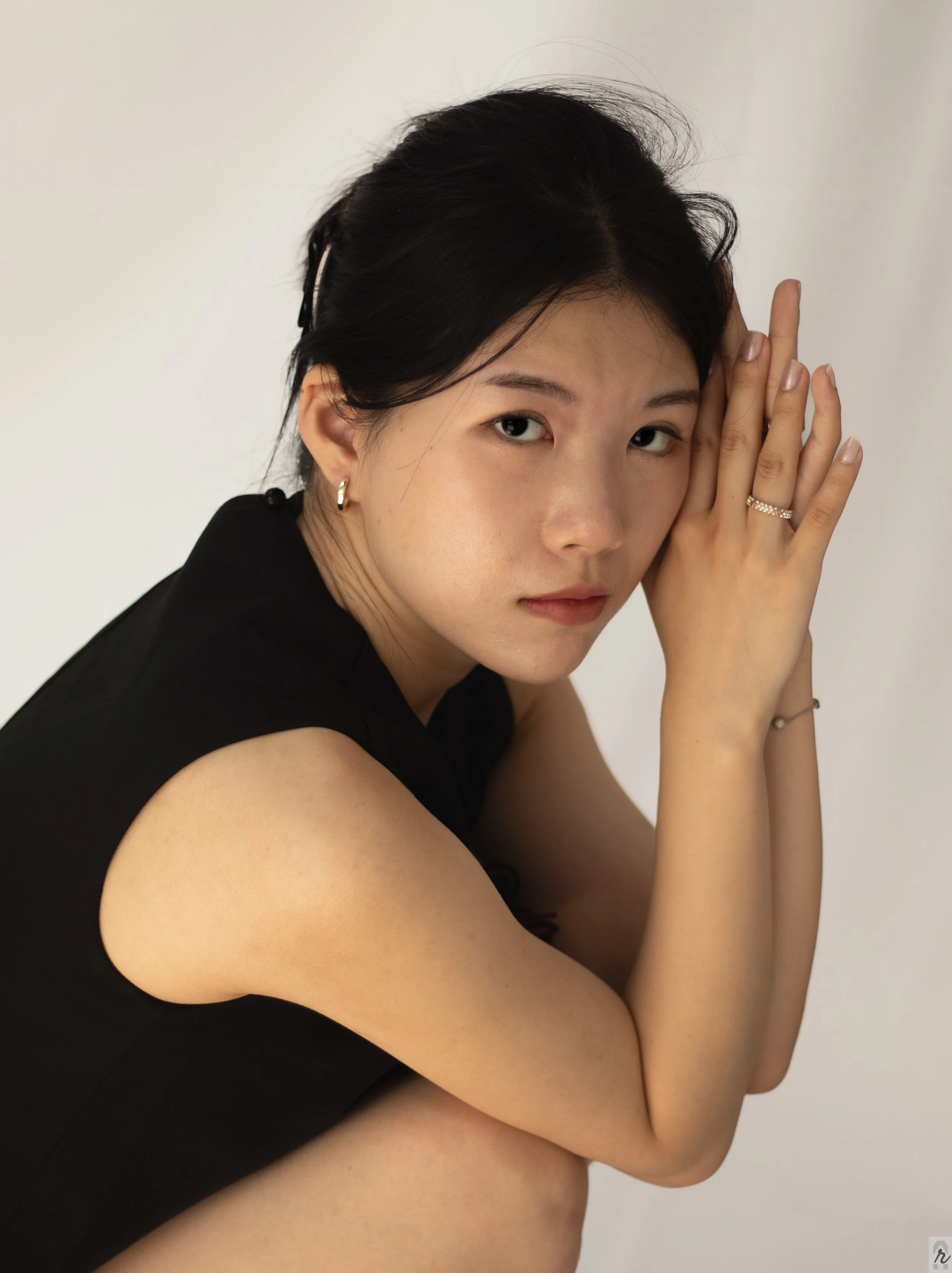 A woman with dark hair and light skin, wearing a black sleeveless top and hoop earrings, is posing with her hands near her face against a plain white background.