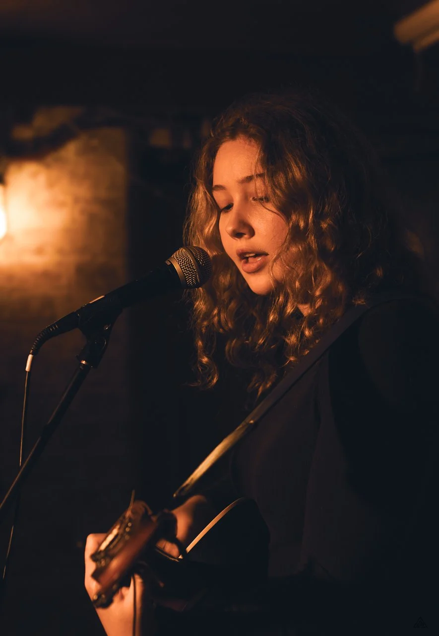 Young woman singing into a microphone while playing an acoustic guitar in a dimly lit setting. Rose & Crown Bar, Kentish Town. London. Camden Songwriting Circle.