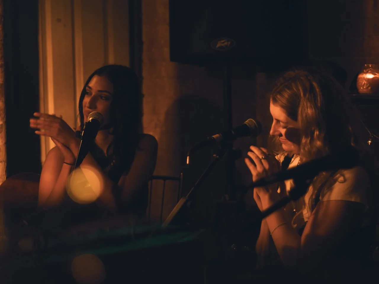 Two women sitting at microphones in a dimly lit setting, possibly performing or speaking, with warm lighting and a brick wall in the background.