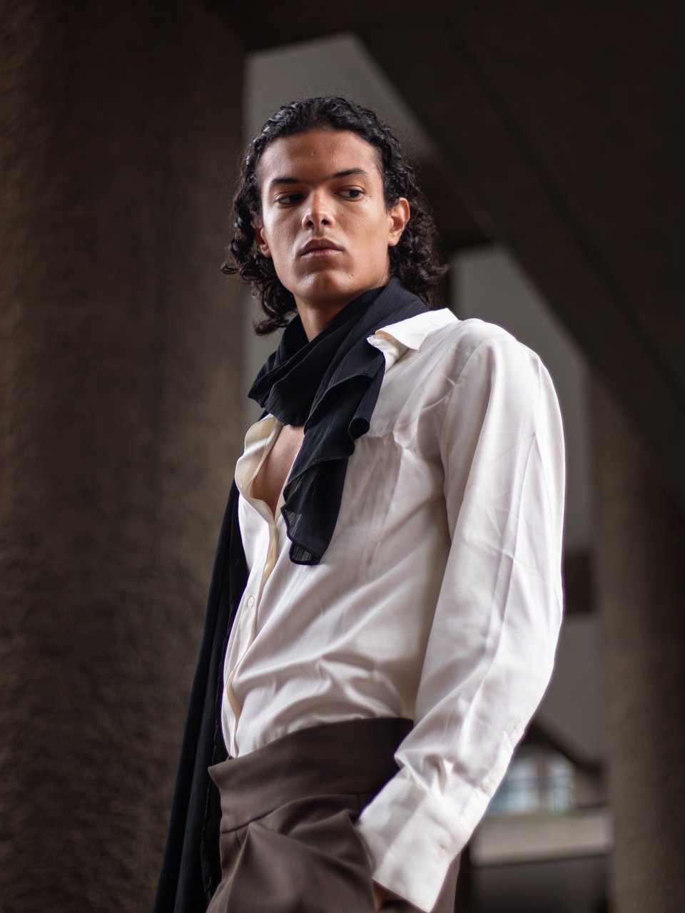 A young man with dark curly hair, wearing a white dress shirt with the top buttons open, a black scarf around his neck, and brown pants, looking sideways down into the camera lens for fashion/ portrait photography. Barbican, London.