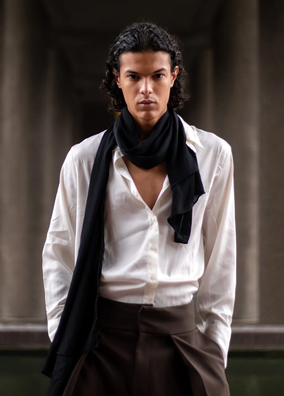 A young man with dark, curly hair and a serious expression, wearing a white dress shirt, black scarf, and dark pants, standing in front of a blurred stone architectural background. Barbican, London. Fashion Photography.