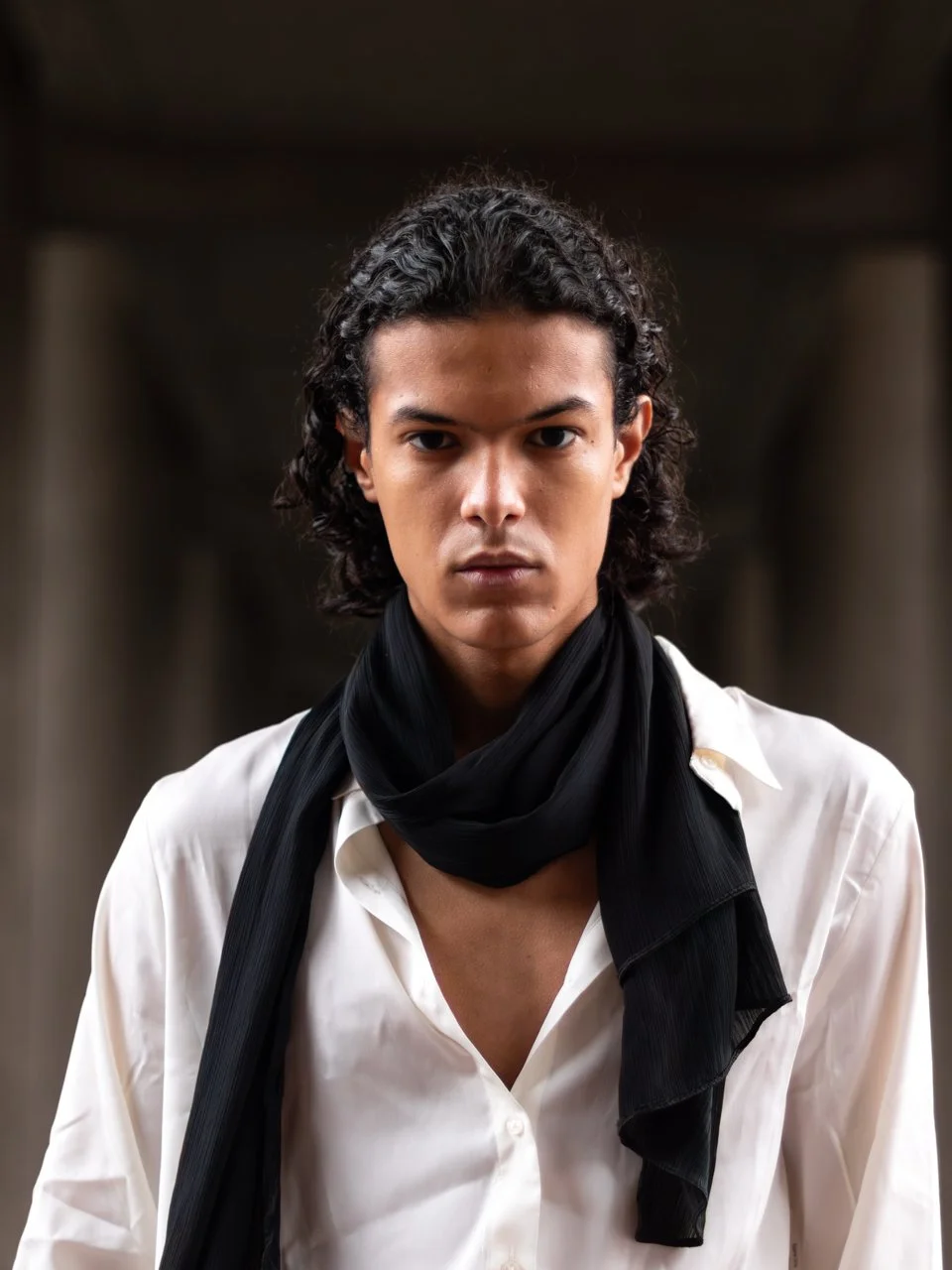 A young man with curly black hair, wearing a white shirt and a black scarf, looking directly at the camera with a serious expression. Outdoor Portrait. Headshot of a young man wearing a white shirt with a black scarf. Barbican, London