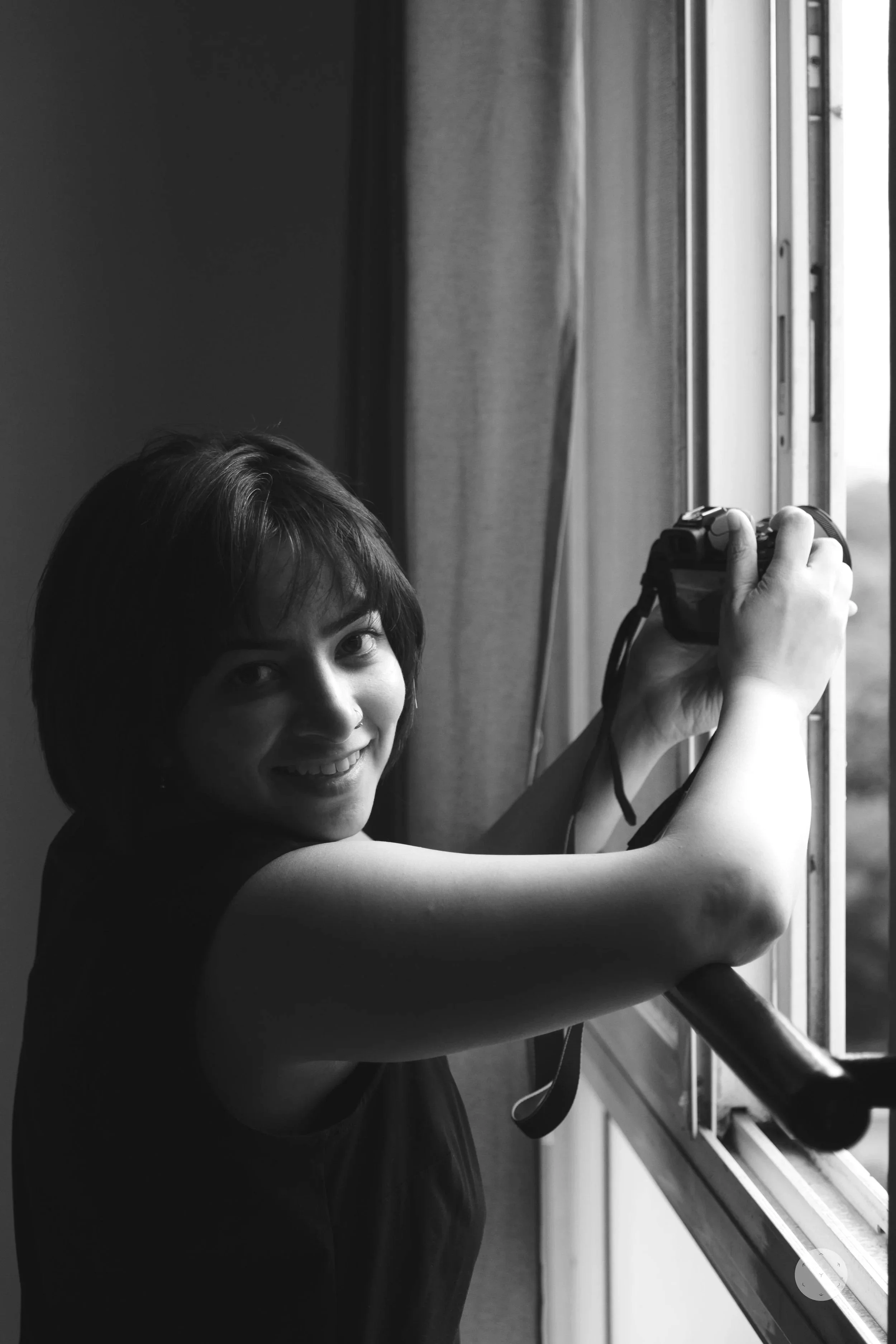 A woman with dark hair is looking at the camera while holding a camera near a window.