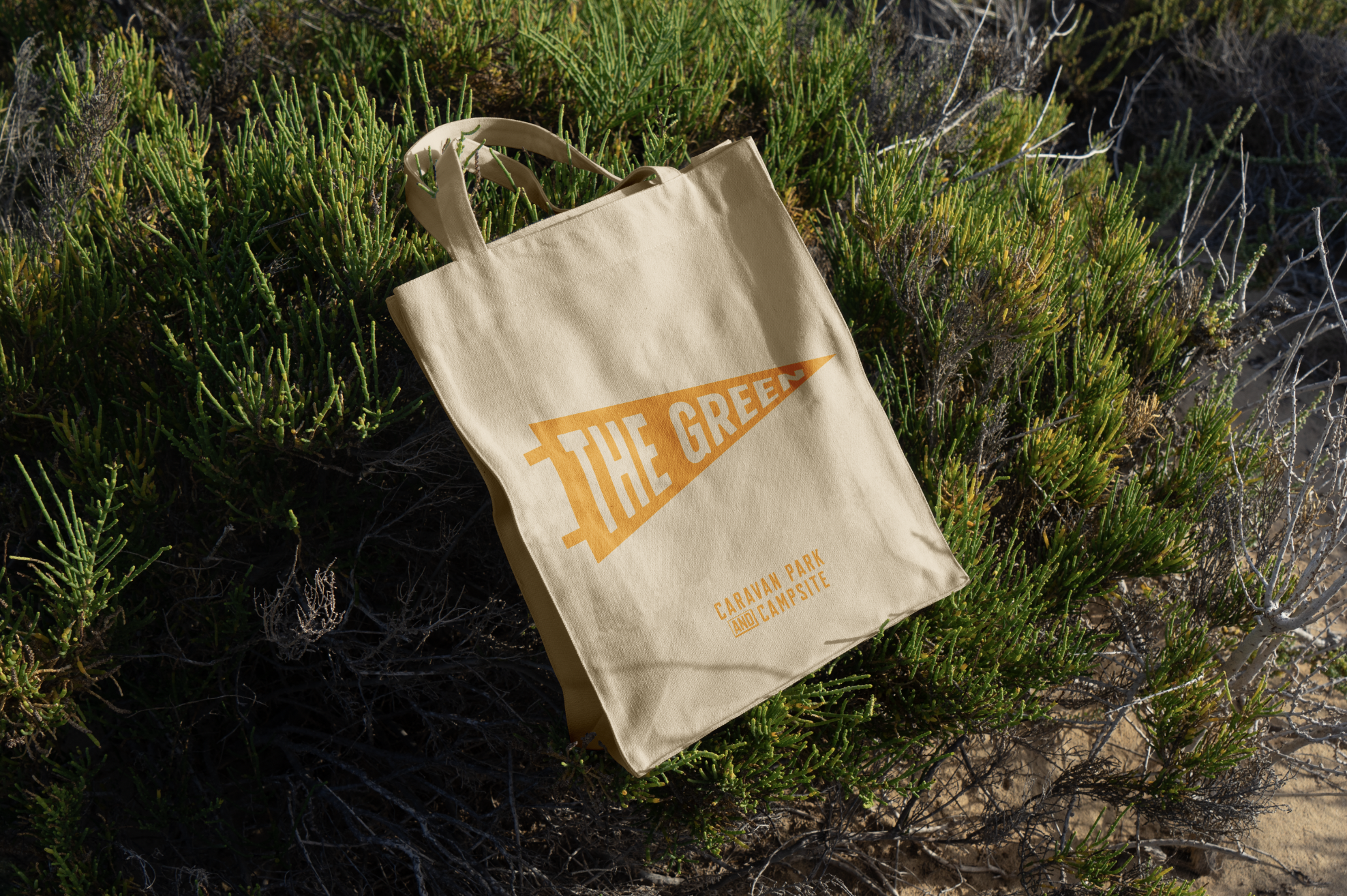 A beige tote bag lying on a patch of green shrubs in natural sunlight. The bag has orange text that reads "THE GREEN" and additional smaller text about Caravan Park and Campsite.