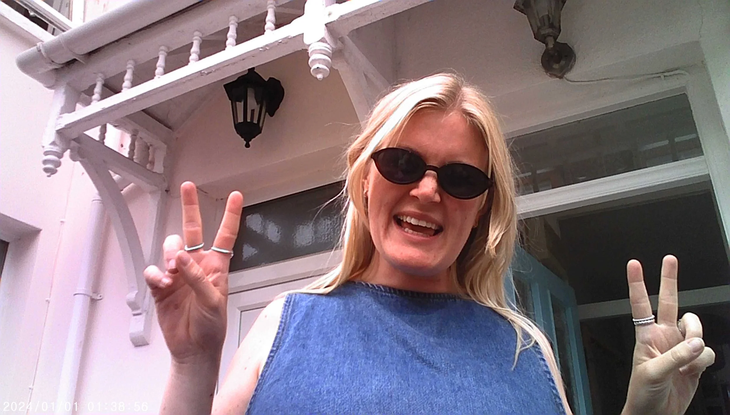 A woman with blonde hair wearing sunglasses smiling and making peace signs with both hands, standing outside a house with white siding, a white porch railing, and a black outdoor wall lantern.