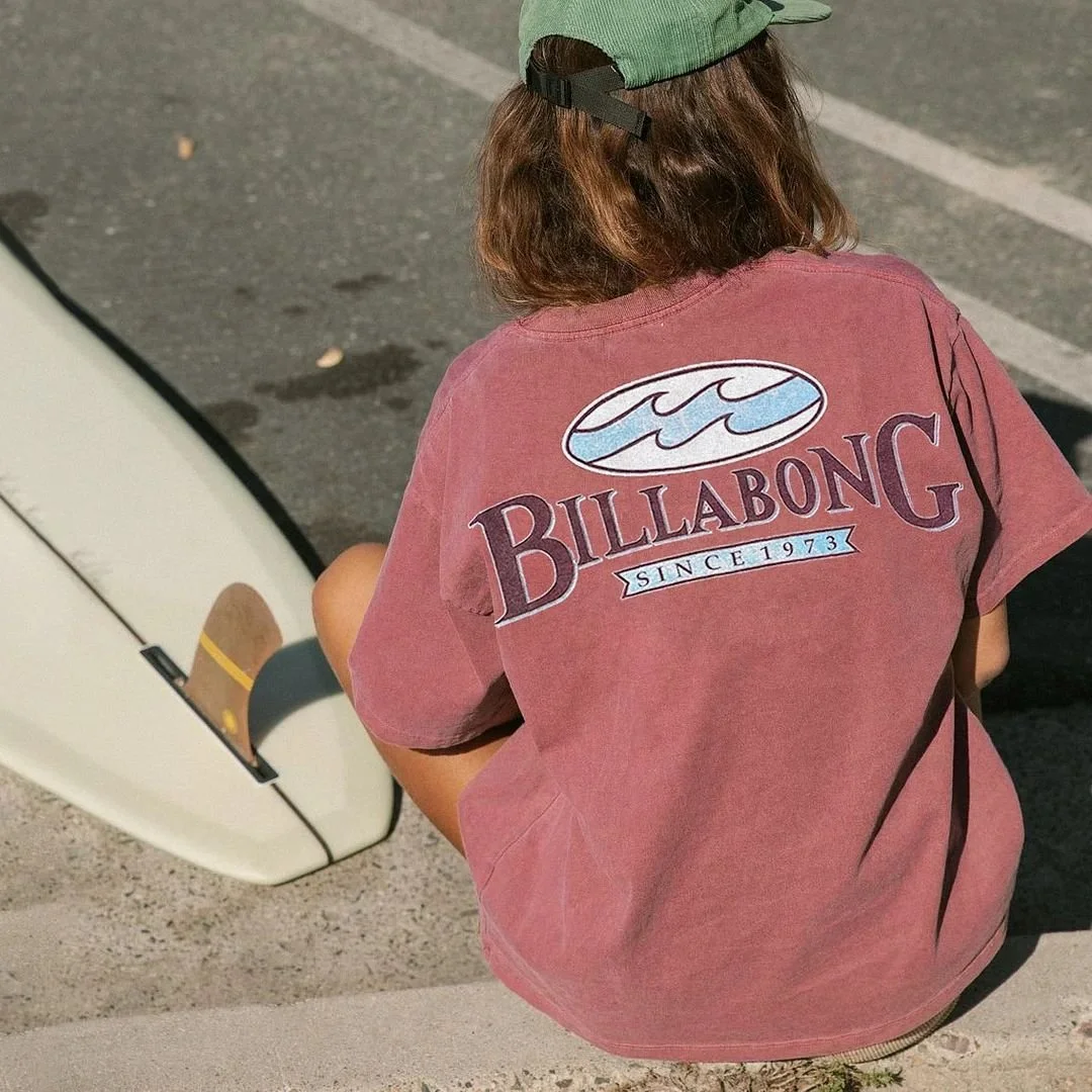 Person in a pink Billabong t-shirt and green cap sitting on curb with skateboard nearby on street.