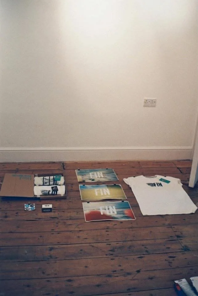 Disassembled or new logo or branding materials with the word 'FIN' on posters and a t-shirt, laid out on a wooden floor in front of a blank white wall.