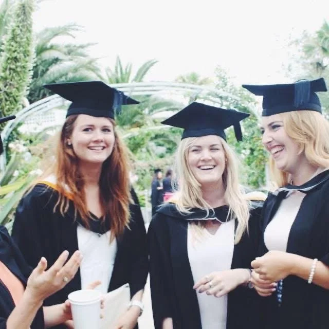 Three women in graduation caps and gowns smiling and celebrating outdoors.
