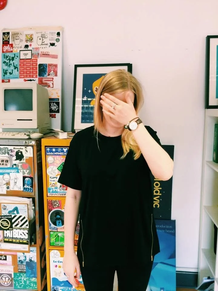 Woman with blonde hair wearing a black T-shirt and watch, covering her face with her hand, in a room filled with colorful posters and books.