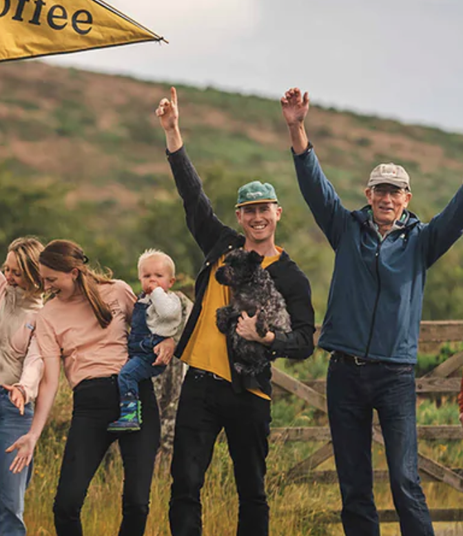 Group of people celebrating outdoors with a man holding a small black dog, smiling and raising their hands, on a grassy hillside with a wooden fence and cloudy sky.