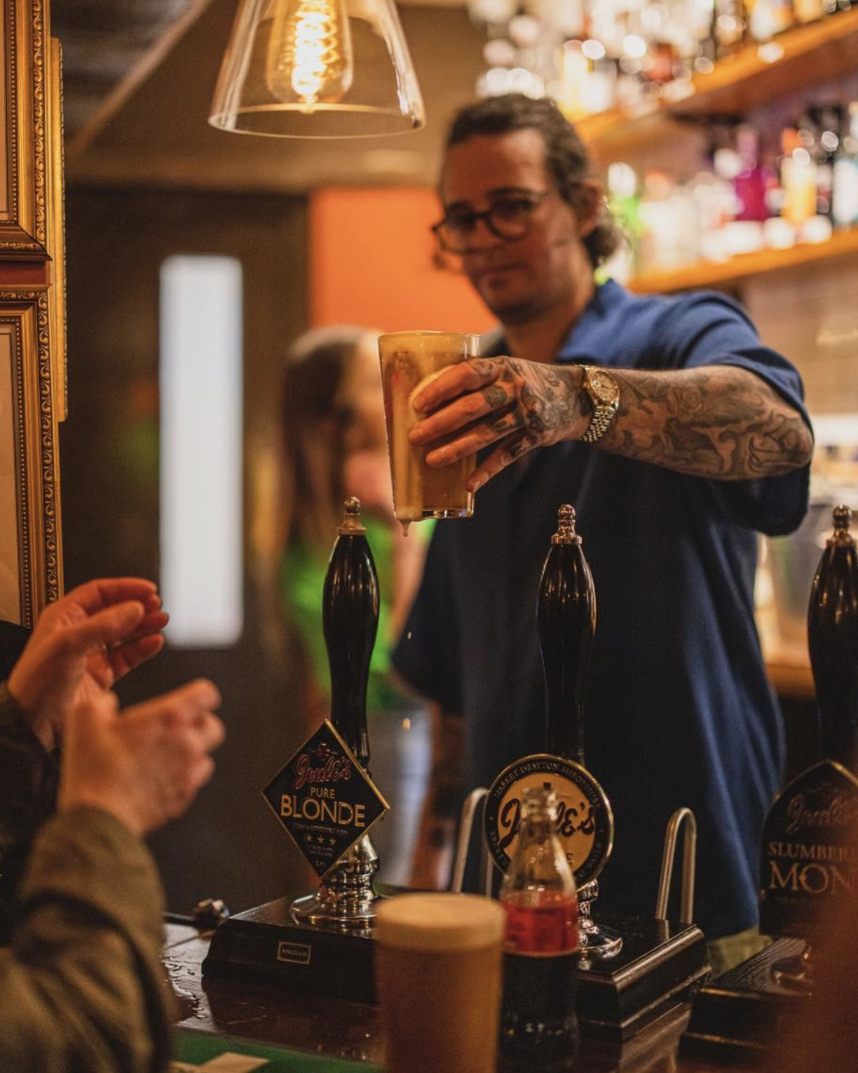 A bartender with tattoos and glasses is pouring a beer from a tap at a bar. The bar area has multiple beer taps and bottles on shelves in the background. A person in the foreground is reaching out.