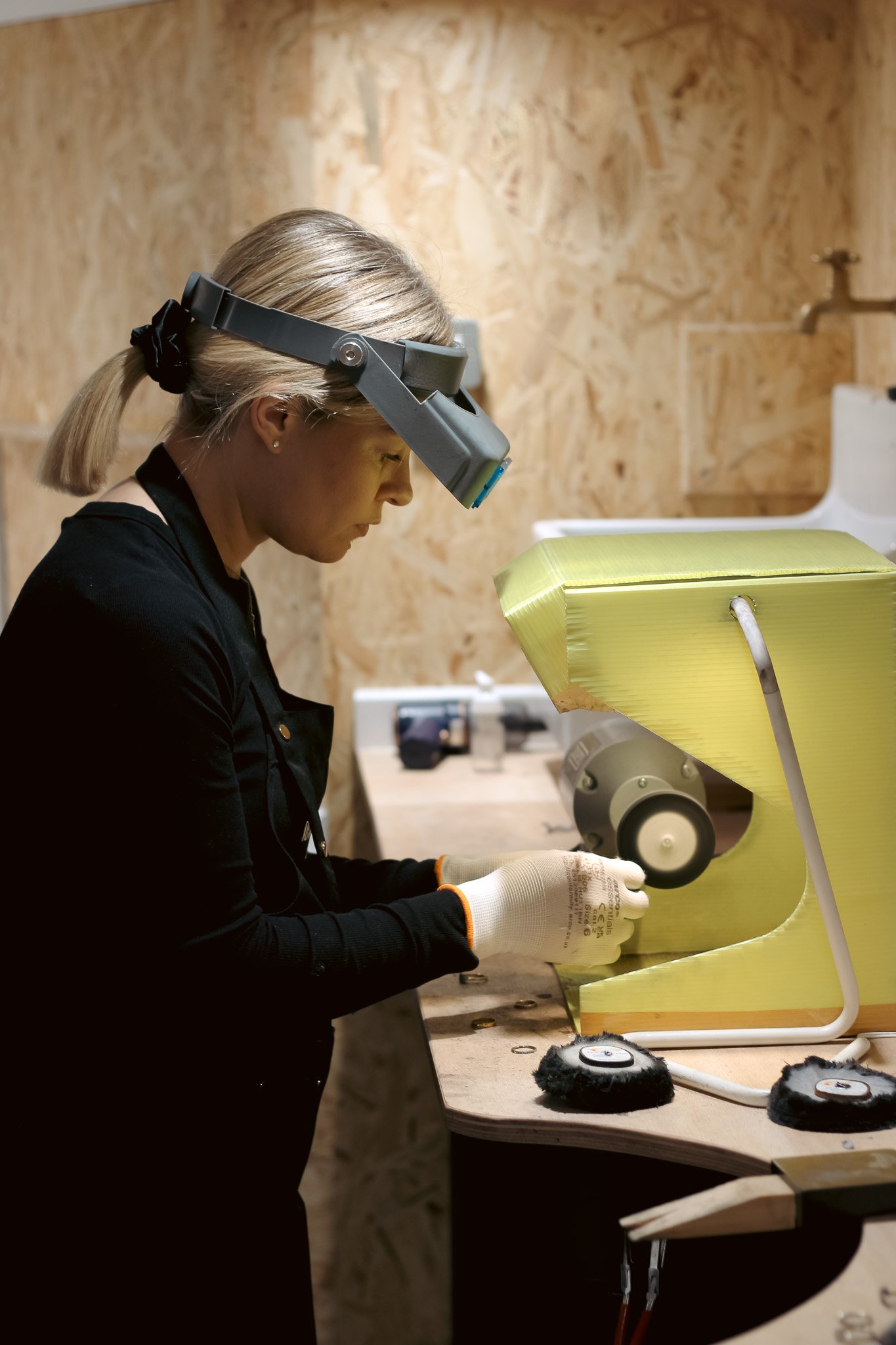 A woman working in a workshop wearing a headlamp and gloves, assembling or repairing a yellow chair with a white frame.