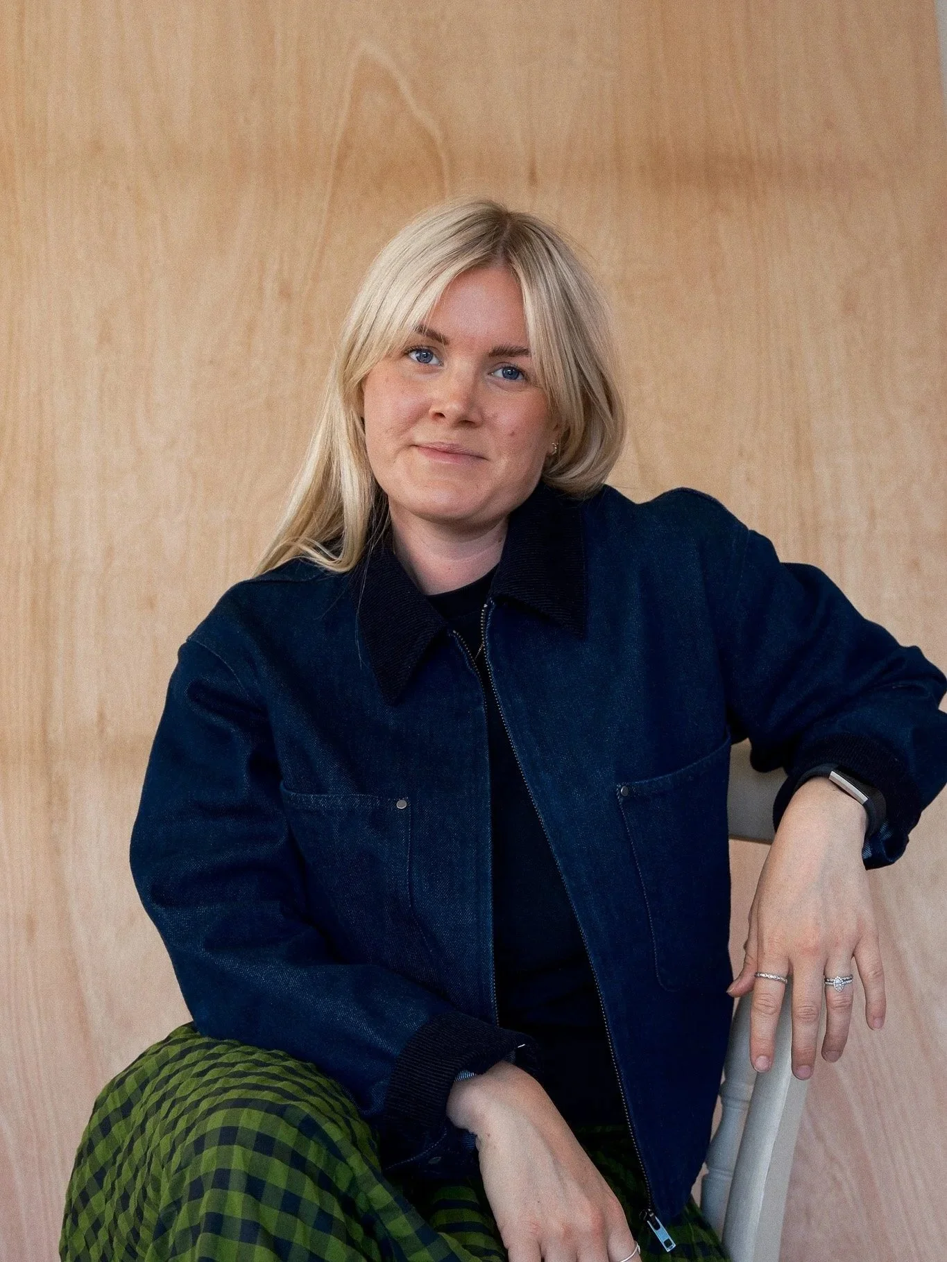 A woman with blonde hair, blue eyes, wearing a dark denim jacket, sitting on a chair against a wooden wall.