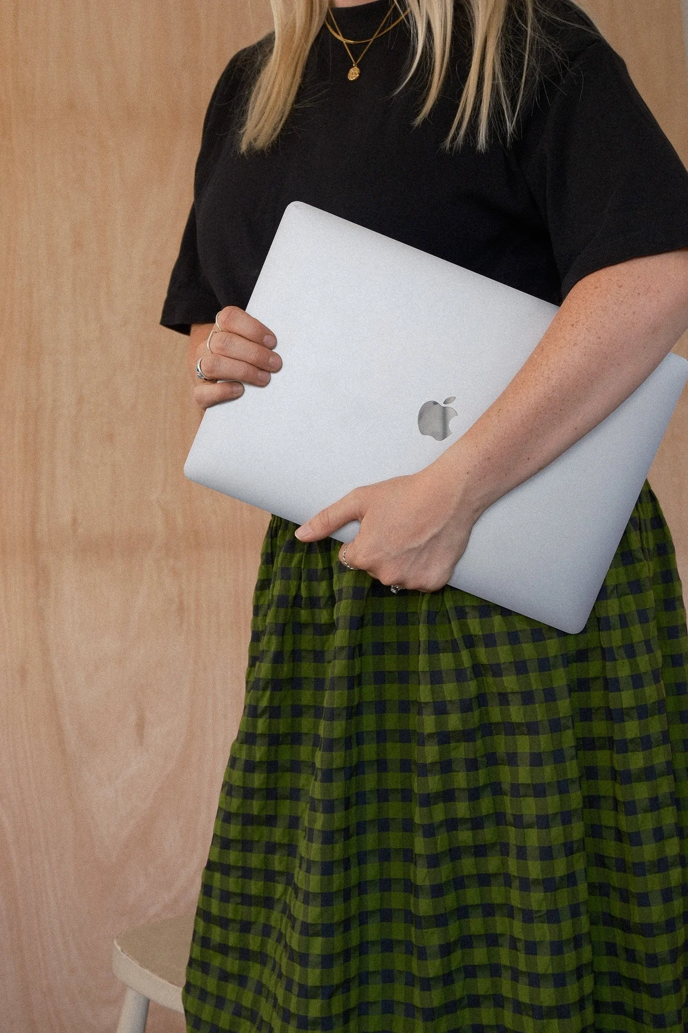 A woman holding a silver MacBook laptop with a wooden background and a chair visible in the scene.