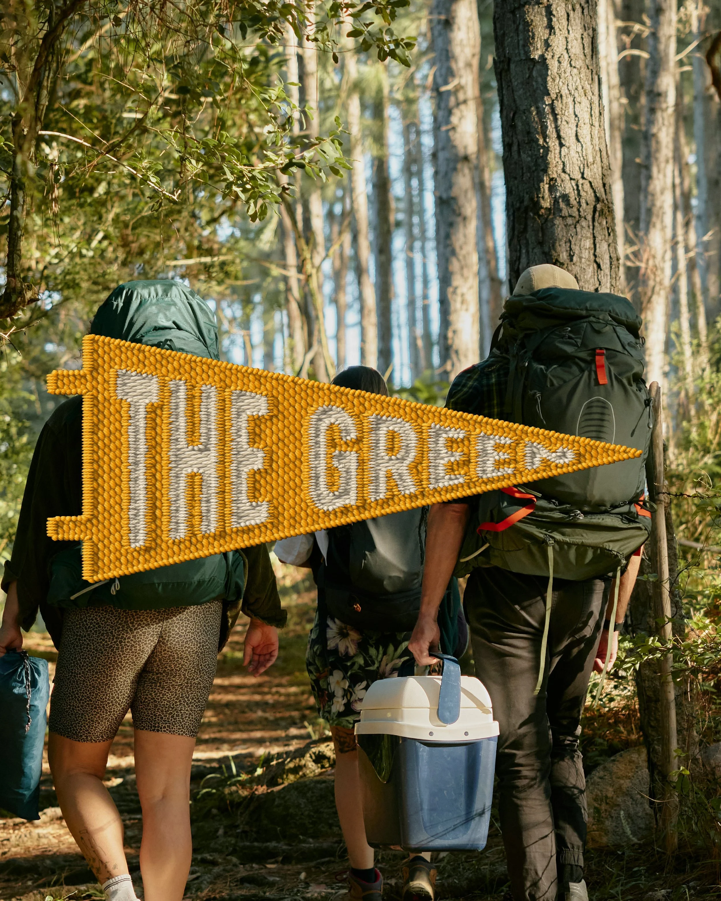 A group of people walking in a forest, carrying backpacks and outdoor gear, with one person holding a cooler. A partially visible yellow sign with white and orange text is in the foreground.
