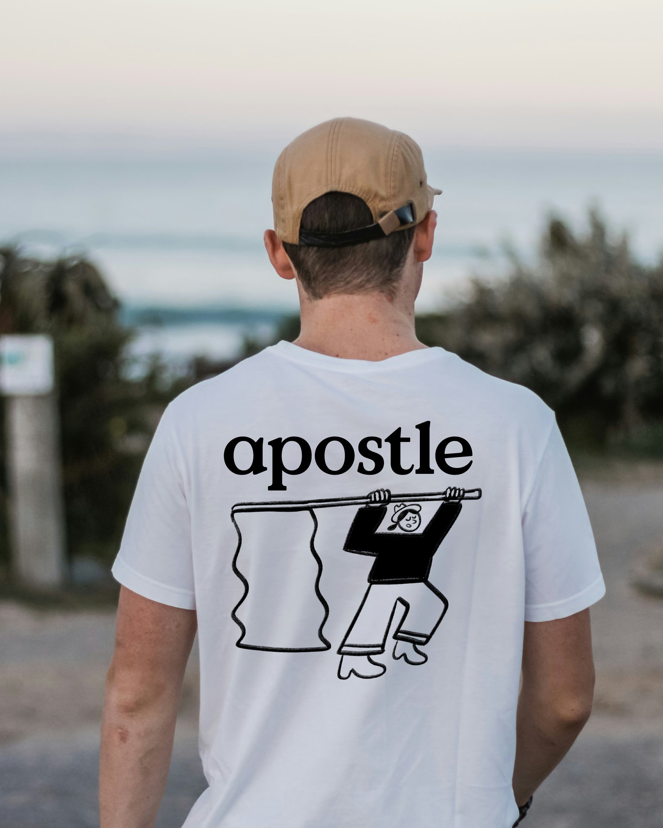 A person wearing a beige cap and a white t-shirt with a black and white graphic of two coffee cups labeled 'a postle' standing outdoors near the ocean with trees and sky in the background.
