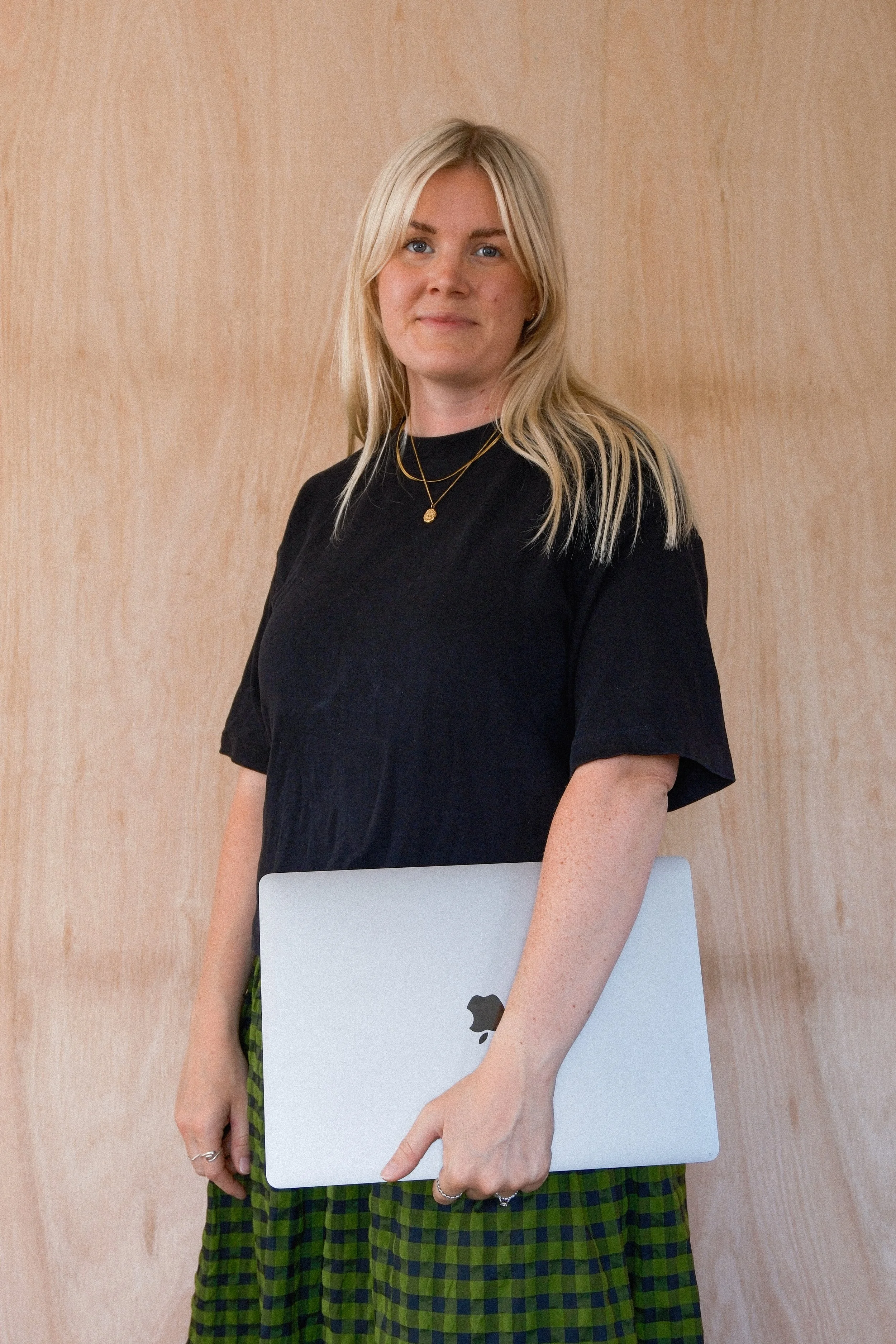A woman with blonde hair wearing a black top and green plaid skirt holding a silver laptop, standing against a wooden wall.