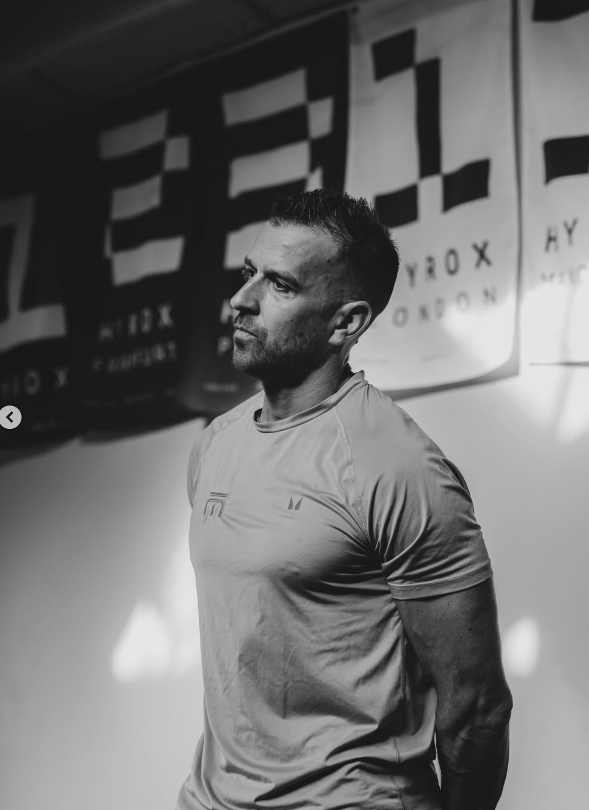 A man in a light-colored athletic shirt standing in a gym or training room with a focused expression, with banners hanging in the background.