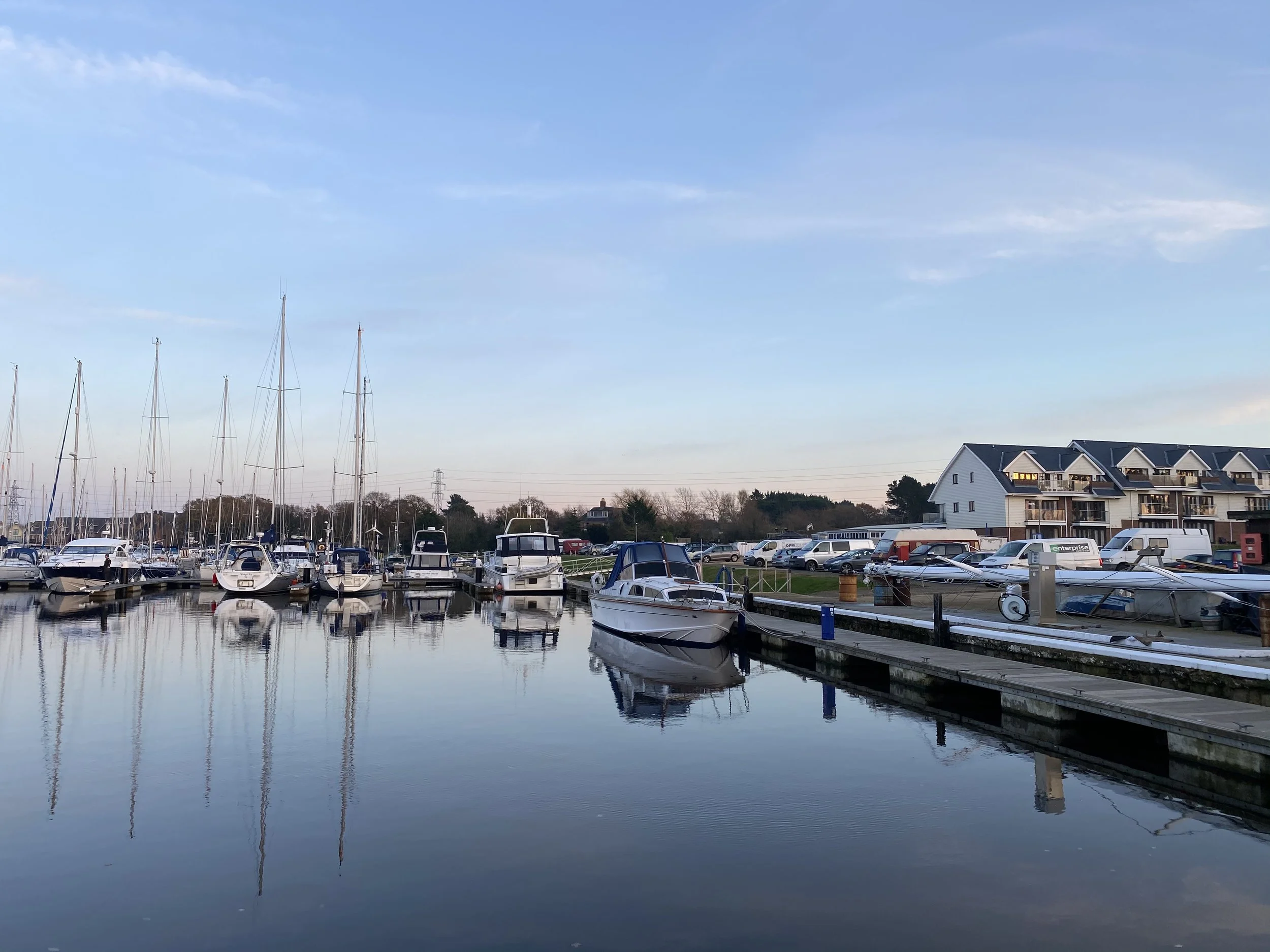 View of a marina with sailboats and motorboats docked along a pier, calm water reflecting the boats, residential buildings in the background, partly cloudy sky.