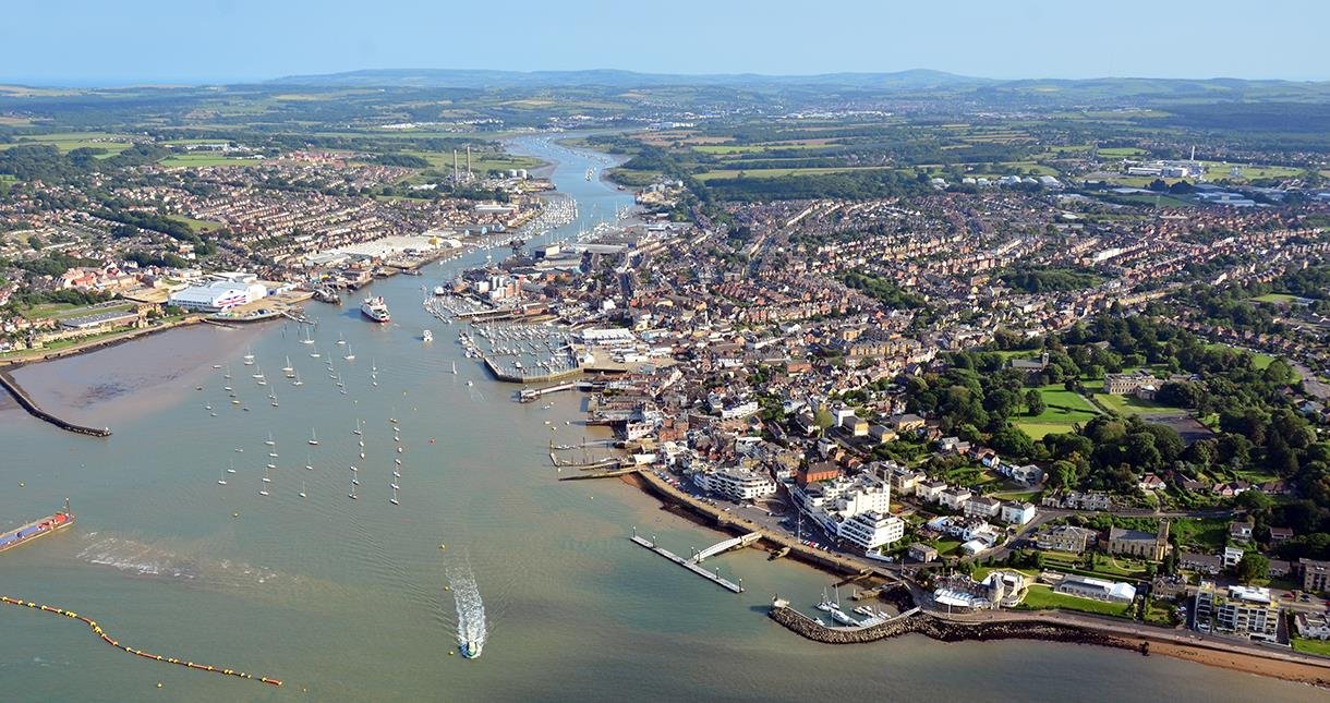 Aerial view of a coastal city with a harbor filled with boats, surrounded by residential and commercial buildings, green parks, and rolling hills in the background.