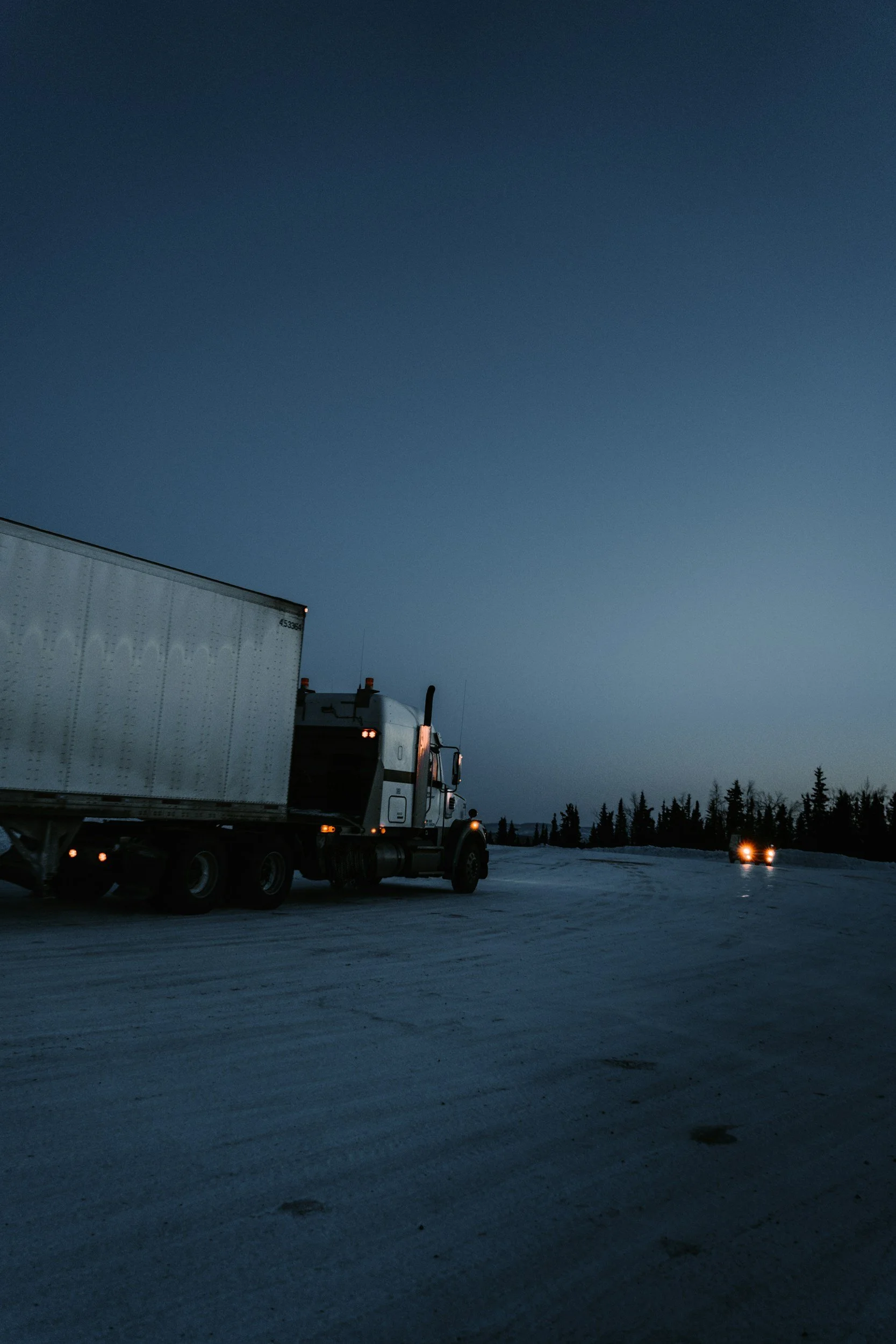 Dark winter evening with a semi-truck in the foreground and headlights of another vehicle in the distance on snow-covered road, surrounded by a forest of evergreens.