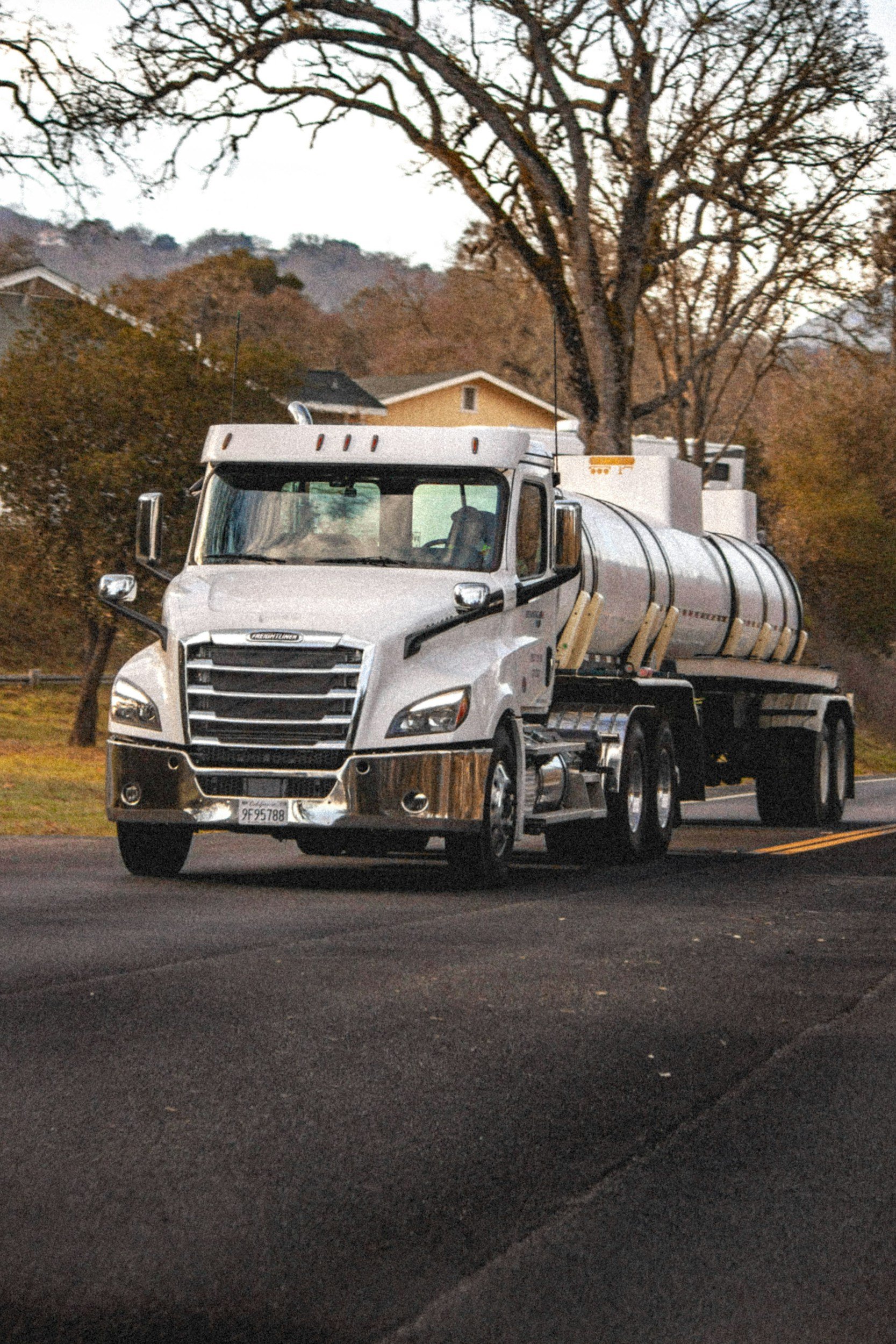 A white fuel tanker truck driving on a paved road through a suburban neighborhood during the daytime. The background features leafless trees and houses with trees on hilly terrain.