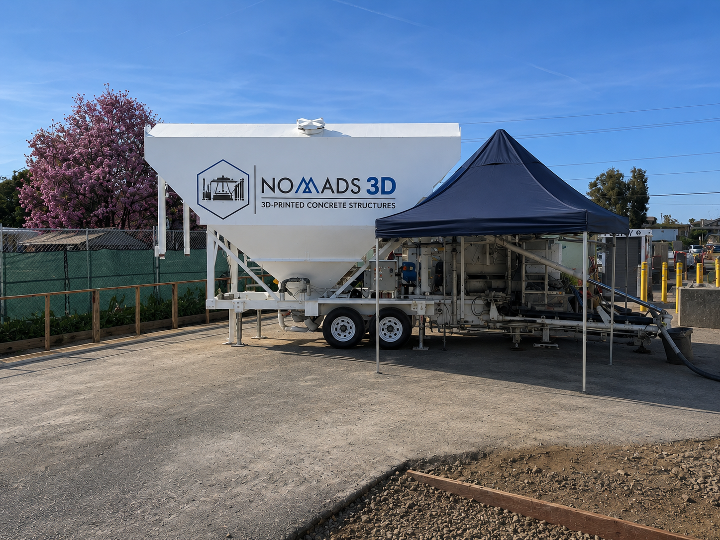 A mobile concrete mixing truck branded NO MADS 3D, with a blue canopy, parked on a construction site with a dirt and asphalt surface, a row of yellow parking barriers, and a background of trees and buildings under a clear blue sky.