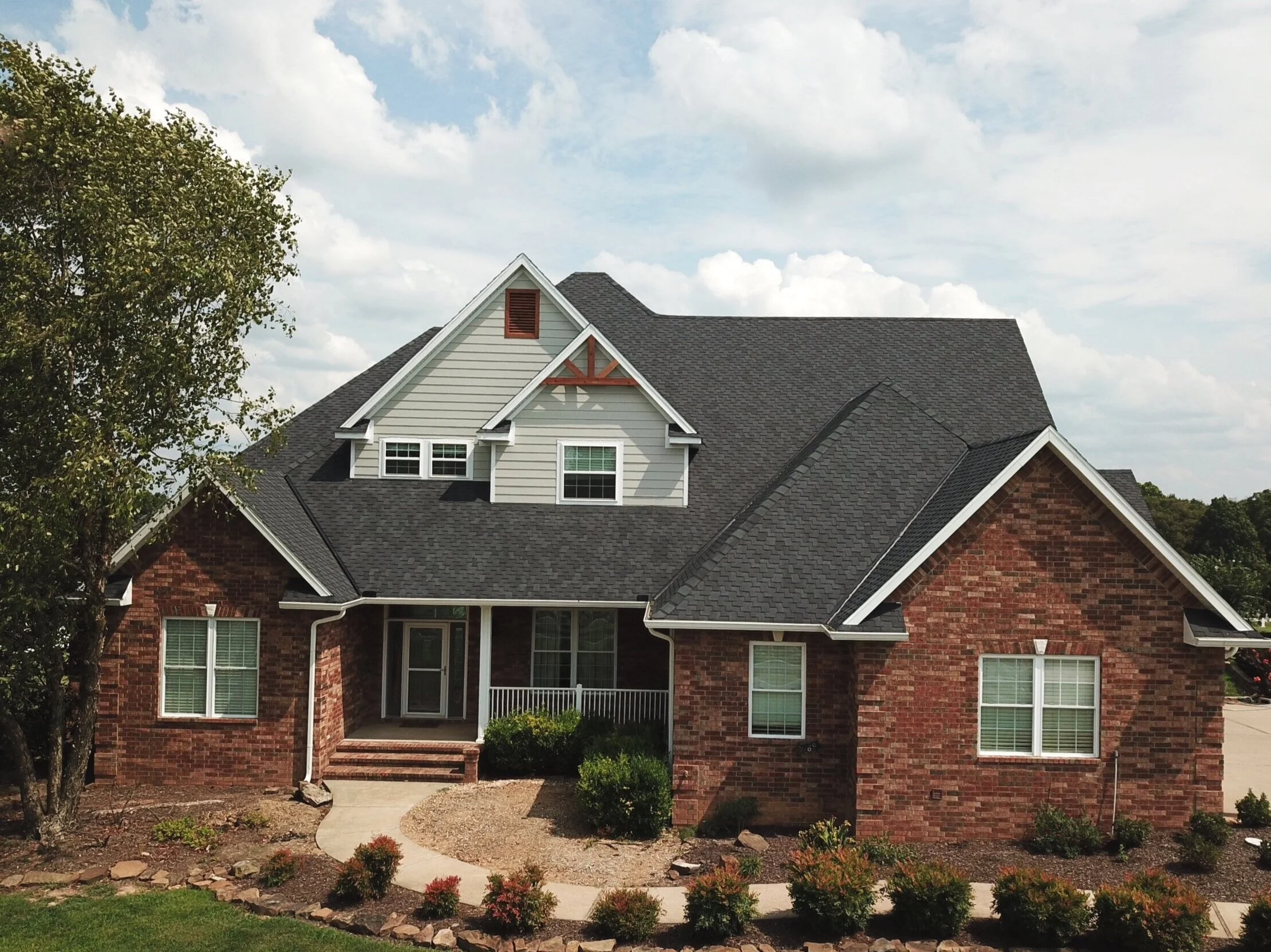 A house with a brick lower level and white upper siding, multiple windows, a small porch, and a gray shingled roof, with a tree to the left and a landscaped front yard.