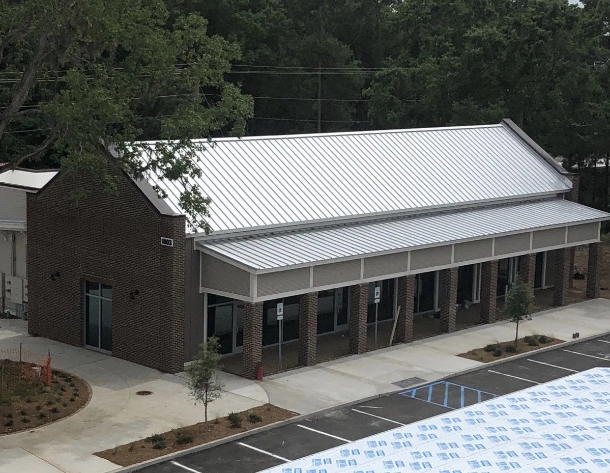 A small commercial building with a modern design, brick exterior, large glass doors, and a metal roof. There are parking spaces and small trees in front.