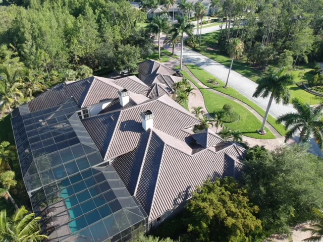 Aerial view of a house with a tiled roof, a solar-paneled screened pool enclosure, and a landscaped yard with palm trees and walking paths.