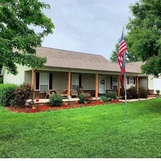 A single-story house with a front porch, an American flag on a pole, green lawn, and trees in the background.