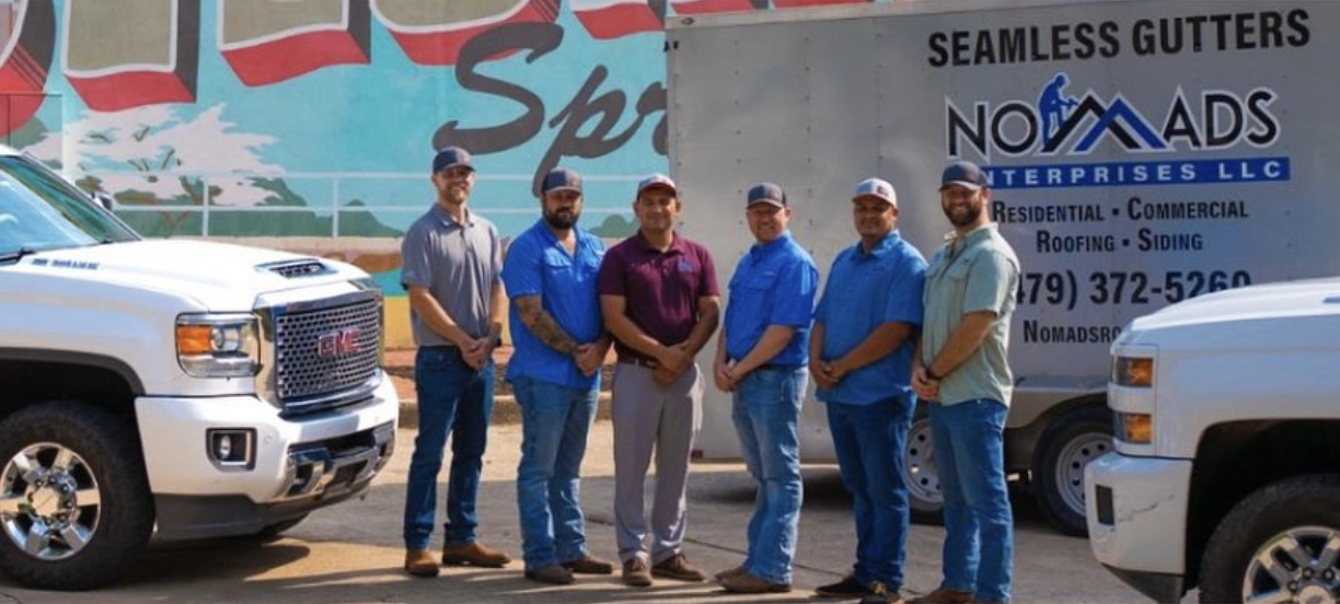 Six men standing in front of a large truck and two white GMC trucks, outdoors, with a mural and a sign for a roofing and siding company.