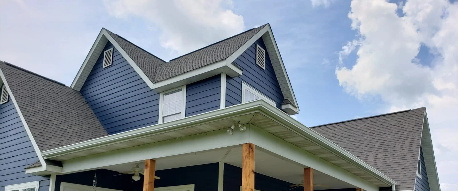 A blue two-story house with white trim and dark gray shingle roof, with a porch supported by wooden posts, under a partly cloudy sky.