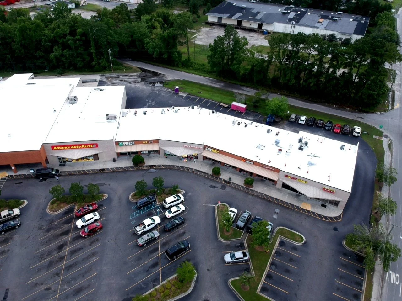 An aerial view of a shopping plaza with parked cars and storefronts, including Advance Auto Parts, Check Cashing, Subway, and Dunkin' donuts. Surrounding the building are trees, parking lots, and a nearby road.
