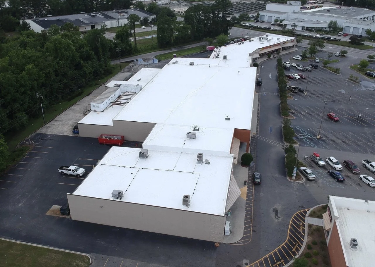 Aerial view of a large shopping mall with a white roof, parking lot with cars, and surrounding trees and buildings.