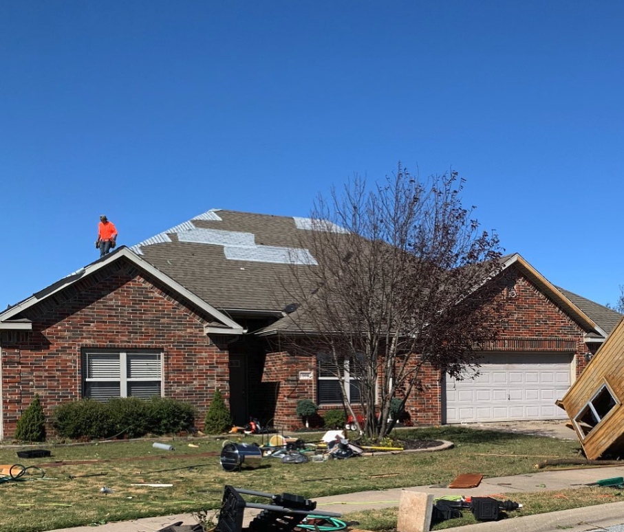 A house with a damaged roof showing missing shingles on a sunny day, with a worker on the roof, and construction tools and equipment scattered on the lawn.