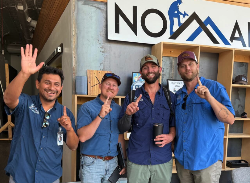 Four men standing together indoors, smiling and pointing upward, wearing blue shirts and caps, with a large sign that reads 'NOVA' in the background.