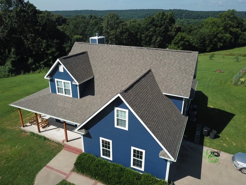 Aerial view of a blue, two-story house with a gabled roof, front porch, and surrounded by green lawn and trees.