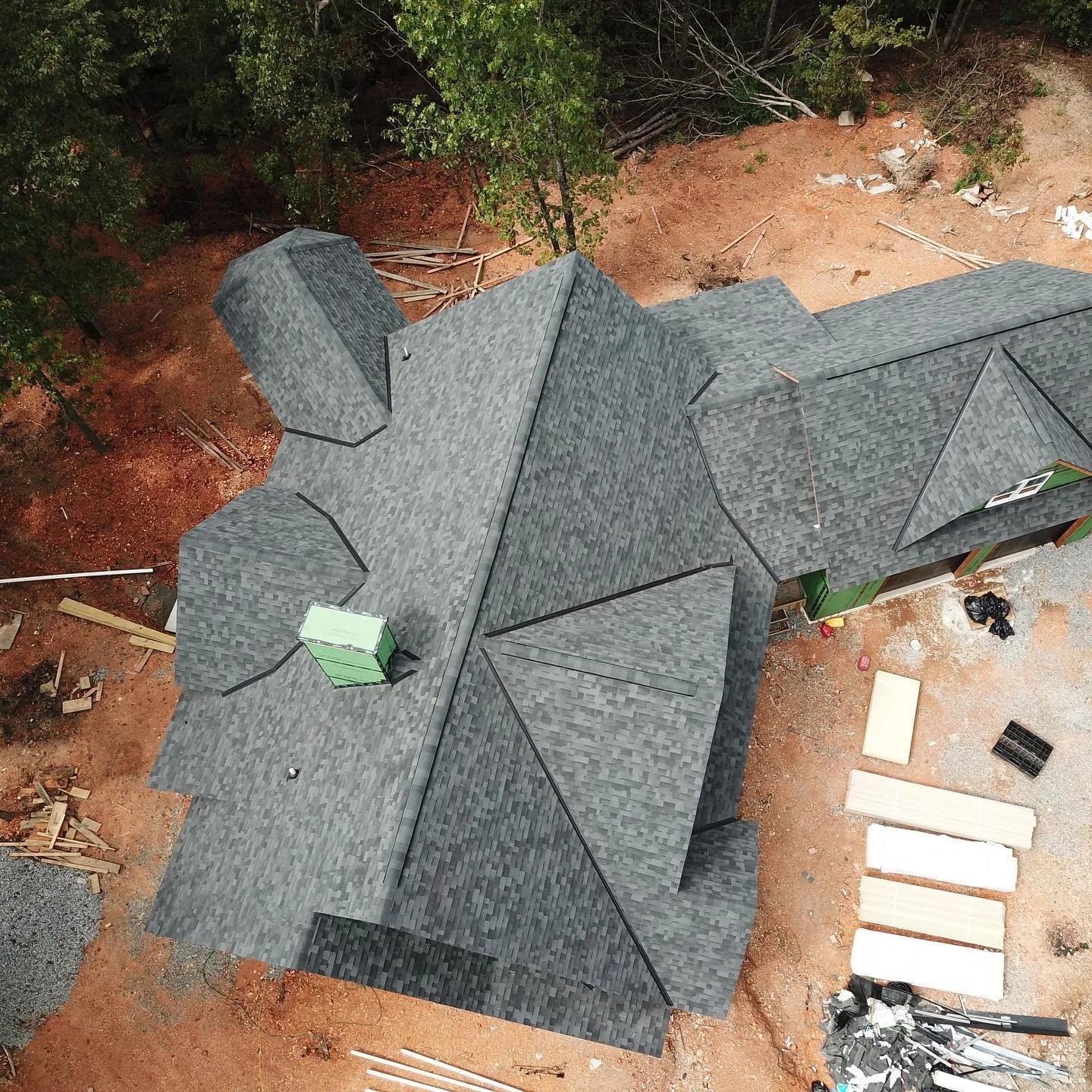 An aerial view of a house under construction with a gray shingled roof surrounded by work materials and earth, with trees at the yard edges.