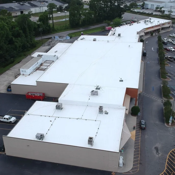 Aerial view of a shopping mall with a large white roof, surrounded by parking lots and some trees.
