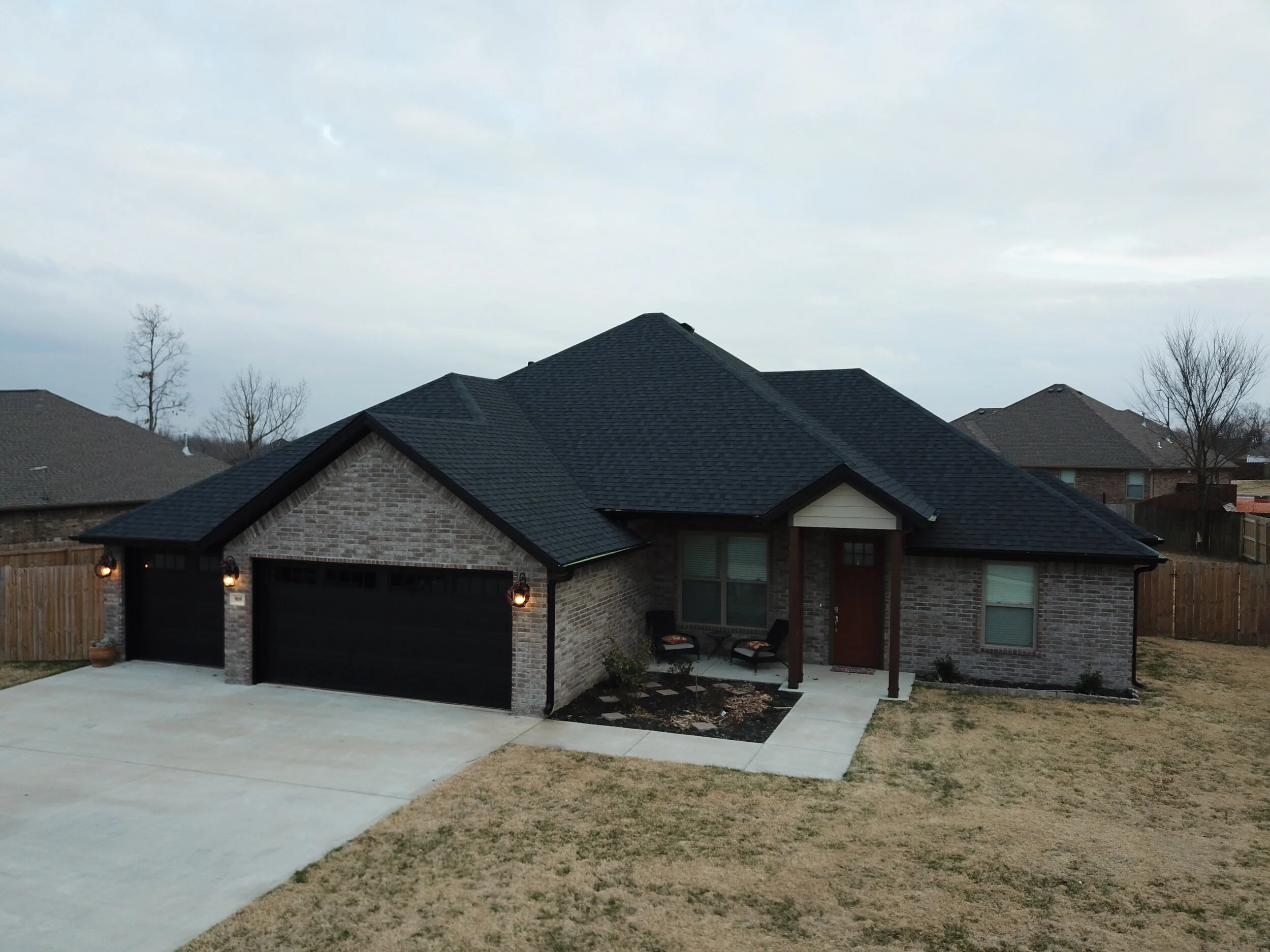 A modern suburban house with a brick exterior, a black garage door, and a dark shingled roof. The driveway is concrete, and there is a small garden with some bushes and outdoor furniture on the porch. The yard has a patchy lawn, and in the background, there are several leafless trees and neighboring houses, all under a cloudy sky.