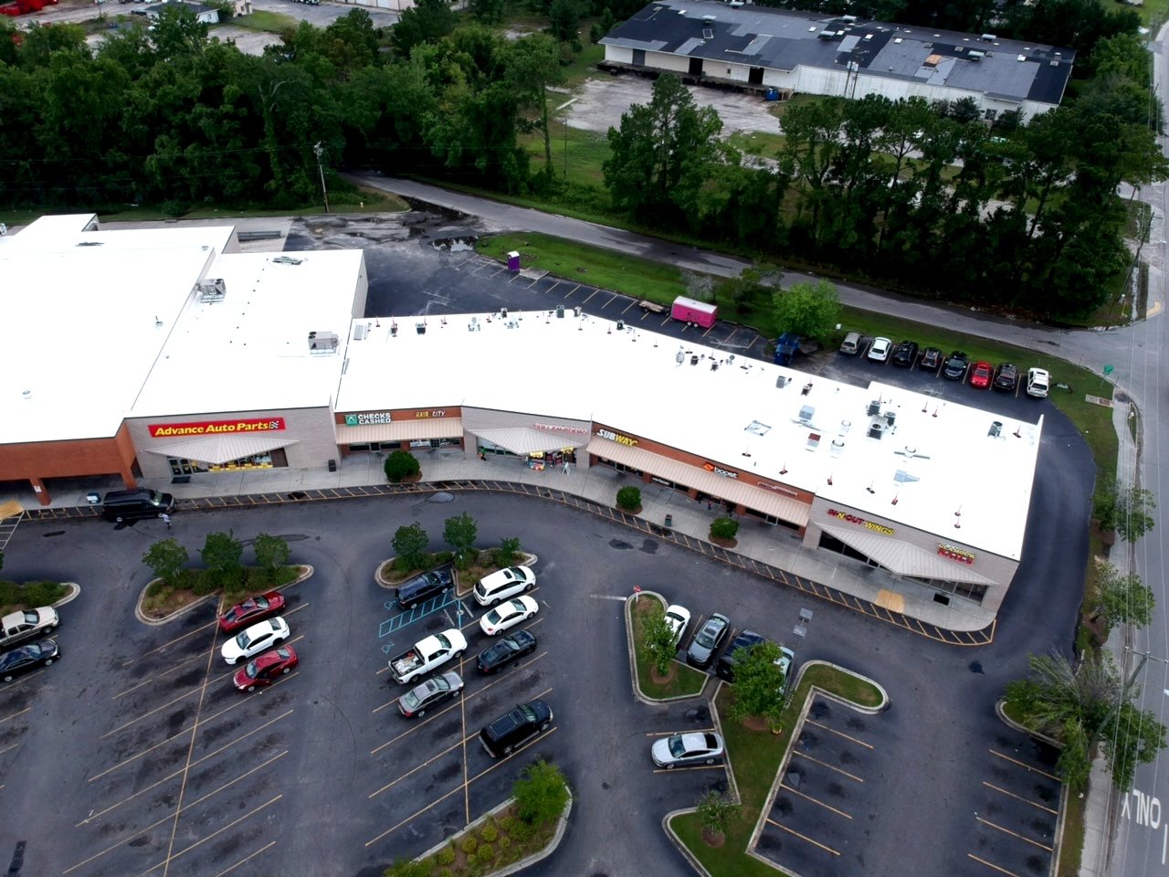 Aerial view of a commercial shopping center with parked cars and storefronts, including Advance Auto Parts, Subway, and other shops. The parking lot has a few empty spaces and is surrounded by trees and a nearby road.