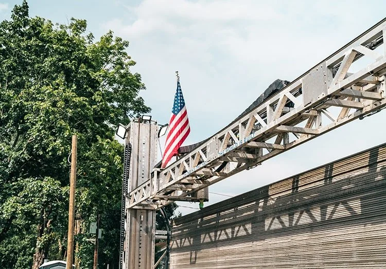 An American flag flying on a fire truck ladder next to a wooden building and green trees.