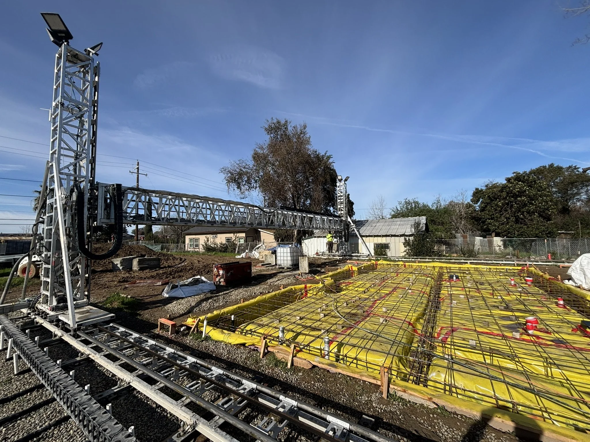 Construction site with steel rebar foundation, yellow tubing, and a tall internal crane against a blue sky, with trees and buildings in the background.