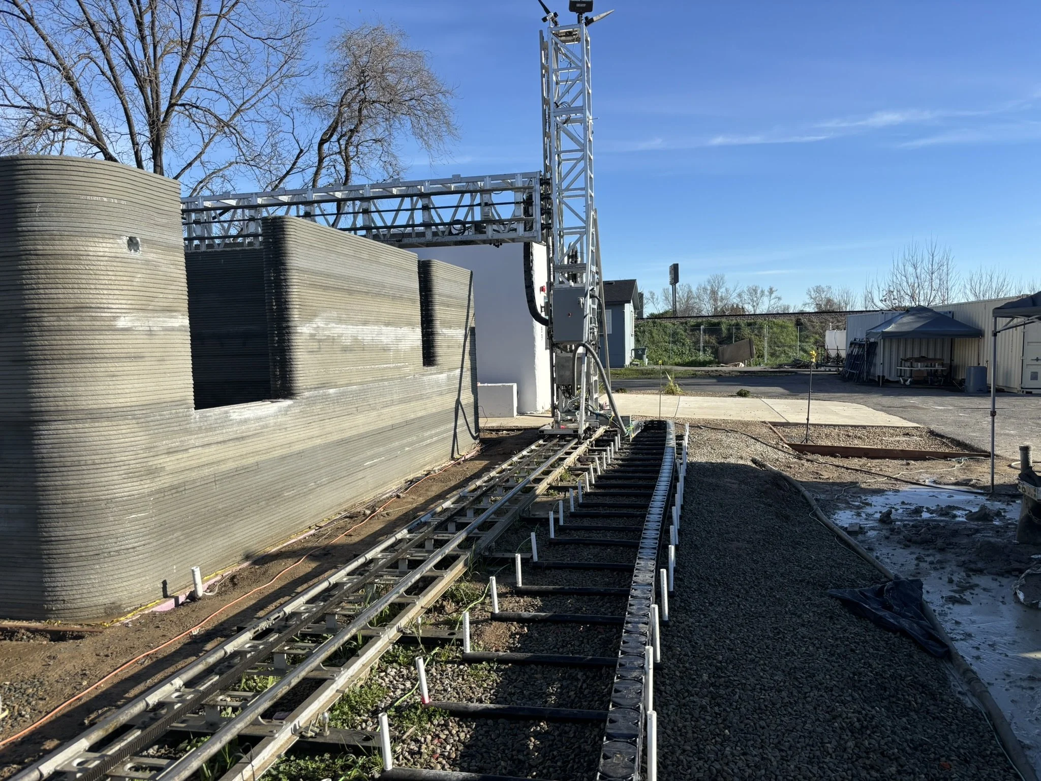 Construction site with a large water tank on the left, metal framework, and a track for machinery. There are trees in the background and clear blue sky.