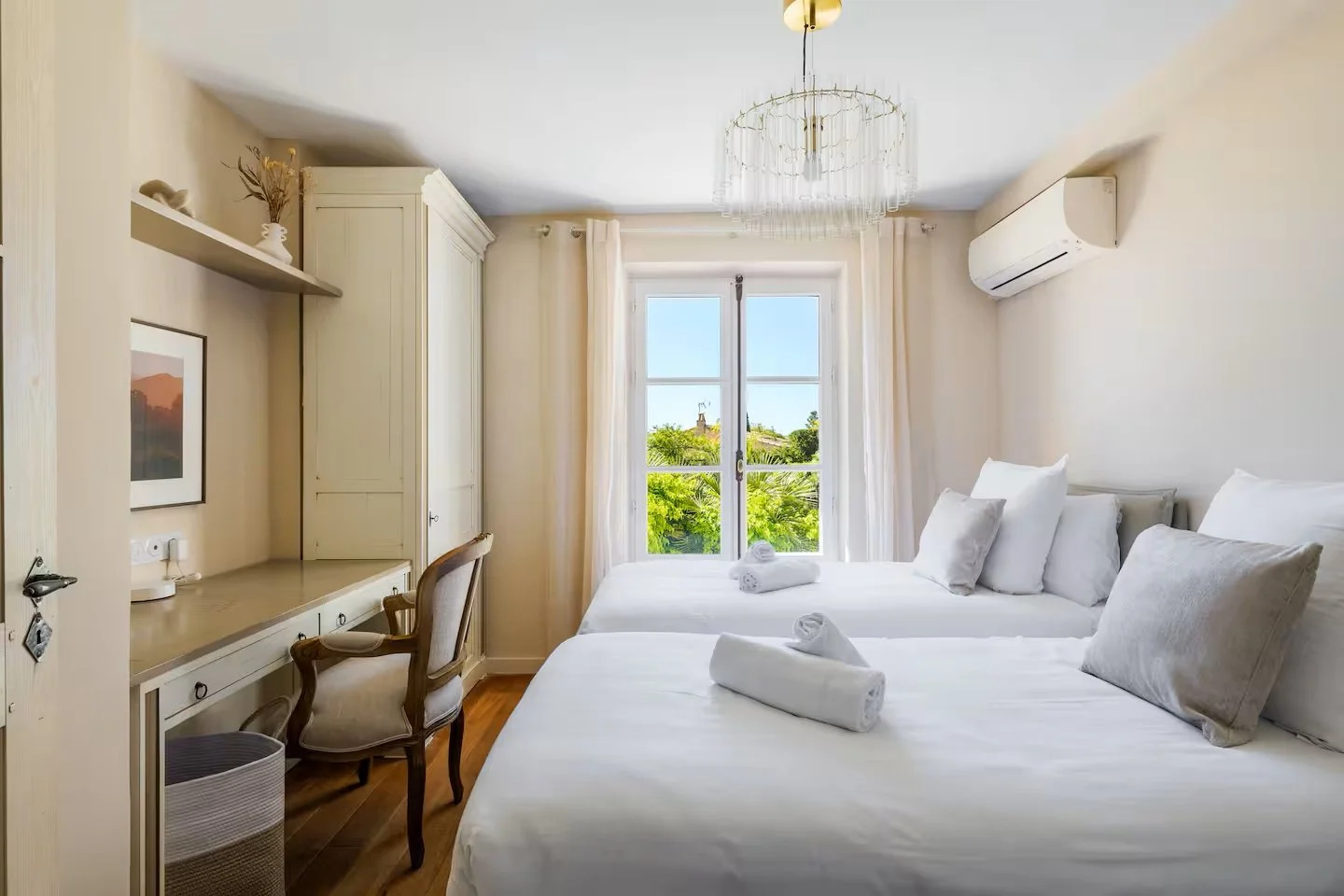 Bright hotel room with two single beds, each with white linens and rolled towels, a window with a view of trees, beige curtains, a wall-mounted air conditioner, a desk with a wooden chair, a framed photo on the wall, and a ceiling chandelier.