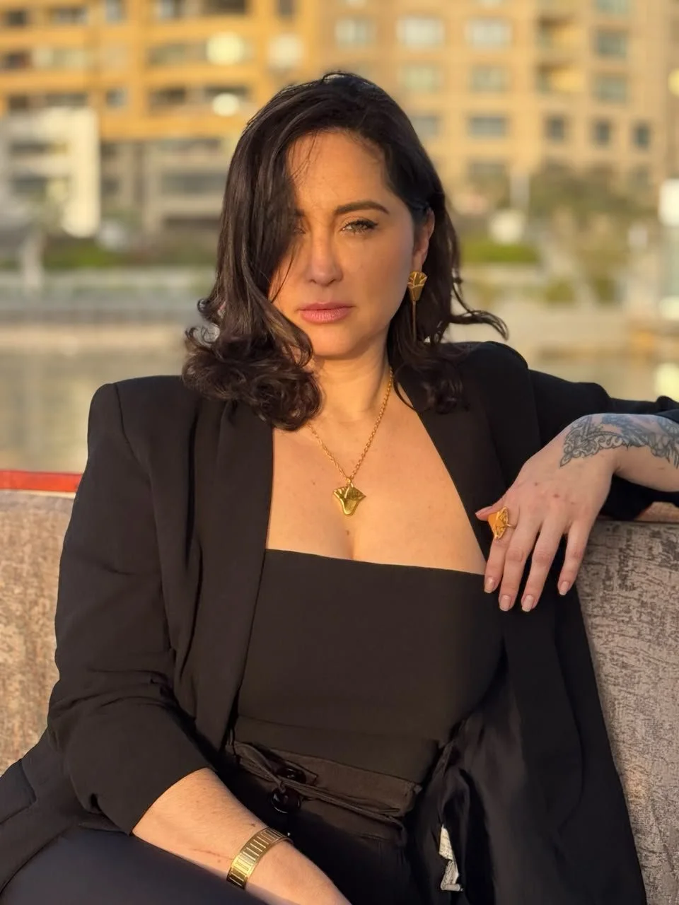 A woman with dark wavy hair, wearing a black strapless top and blazer, sitting outdoors with a cityscape and water in the background, adorned with gold jewelry and a tattoo on her hand.