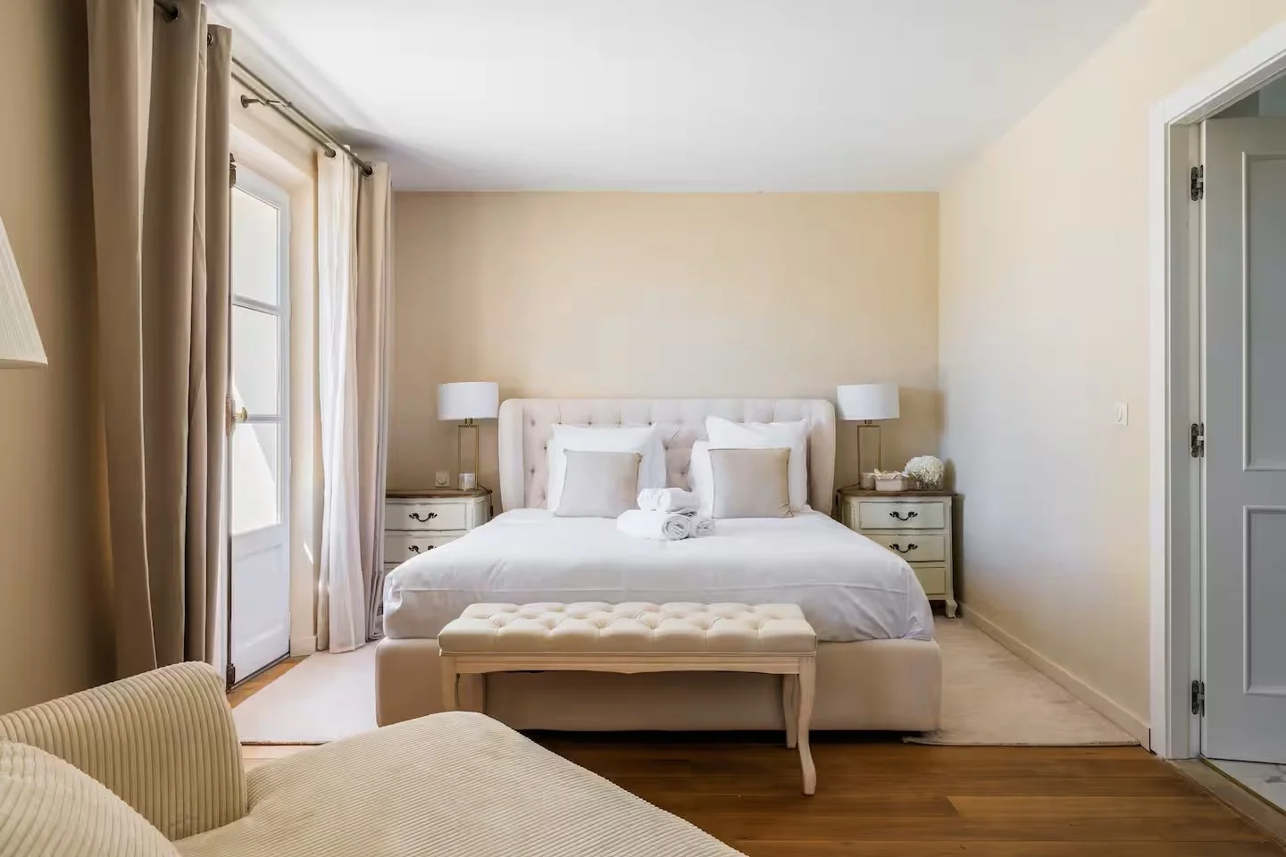 Bright bedroom with a large bed, white bedding, and four pillows, flanked by two nightstands with lamps; a window with curtains, an ottoman at the foot of the bed, and a door on the right.